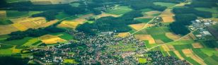 Luftbildaufnahme einer Kleinstadt umringt von grünen und gelben Feldern und Wäldern.