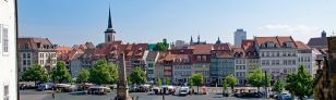 Blick auf den Domplatz von Erfurt. Ein großer Platz mit Oblisk und einigen Marktständen, umringt von alten und neuen Gebäuden, im Hintergund ragt eine Kirchturm hoch.