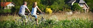 Zwei Personen fahren Fahrrad. Im Vordergrund sind Gräser und Wildblumen zu sehen im Hintergurnd Bäume und Einfamlienhäuser.